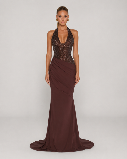 A model wearing a floor-length evening gown with a deep V-neckline, a heavily embellished dark sequined bodice, and a draped satin skirt in a deep burgundy-brown color, set against a plain light gray studio background.